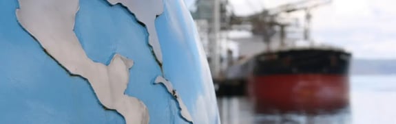 A close-up view of a blue globe sculpture showing parts of North and Central America, with an out-of-focus cargo ship and port equipment in the background.