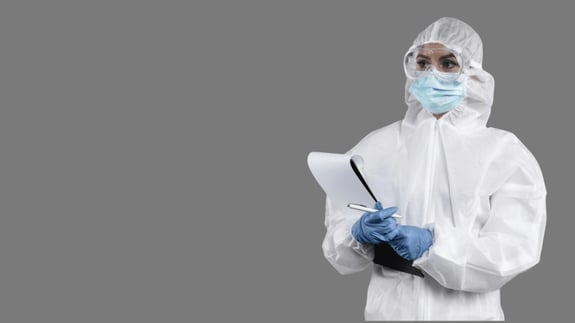 Cleanroom worker in full gown, gloves, and mask holding a clipboard and pen against a grey background.
