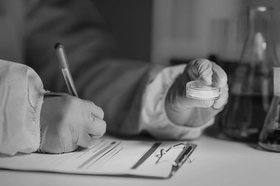 A cleanroom technician wearing gloves writes on a clipboard while holding a petri dish.
